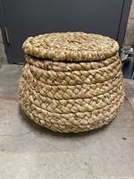 Round handwoven natural wicker storage basket with lid resting on a concrete floor in front of a gray door, showing the basket's shape and lid structure.