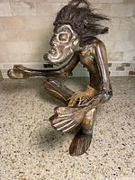 Side view of wooden primitive man figurine seated with arms extended, showing twine skirt and spiky hair made of fibers.