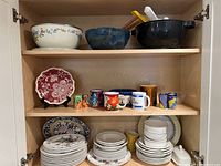 Open cupboard shelves featuring large salad bowls on top shelf with utensils, middle shelf with colorful coffee mugs and a decorative red plate, and bottom shelf stacked with plates, bowls, and cups.