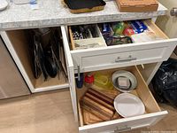 Overview photo showing open cupboard and two drawers with baking trays, flatware, plastic containers, plates, cutting boards, and kitchen wraps.