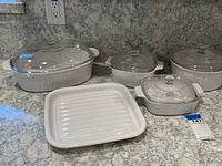 Six covered casseroles and a roasting pan shown on a countertop with marble pattern. Variety of sizes and styles, some unmarked, some marked Corningware with clear glass lids.