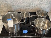 Photo of four stainless steel cookware pieces with glass lids on stovetop, including stockpot, two saucepans, and sauté pan.