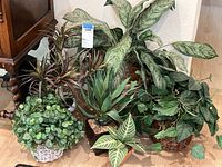 Six faux plants arranged on a floor, showing various sizes, leaf shapes, and pots including woven baskets and plastic pots.