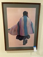 Full view of framed art print showing a Native American figure walking, wrapped in vertical striped blanket, with brown wooden frame and matting.