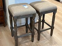 Two wooden kitchen counter stools with tan fabric cushioned seats and decorative nailhead trim, showing overall condition and style.