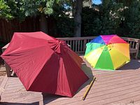 Two umbrellas set on an outdoor wooden deck: the maroon patio umbrella with wooden pole and the rainbow beach umbrella with metal pole.