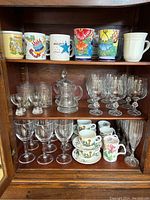 Photo of three shelves showing assorted souvenir mugs on top shelf with colorful prints including Florida, Milpitas, and birthday designs, a plain white mug, small pitcher, clear drinking glasses, and floral patterned cups and saucers on lower shelves.