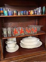 Three shelves of various glassware including souvenir shot glasses featuring city designs, clear glasses with orange floral patterns, and white Mikasa china serving ware