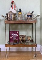 Full view of bar cart with items arranged on both wood shelves showing all accessories, ice buckets, glasses, decanter, soda siphon, and decorative book.