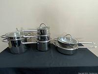 Full set of stainless steel Wolfgang Puck Cafe Collection cookware including stock pot, saucepans, saute pan and pan arranged on black surface.