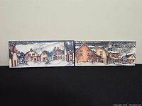 Two winter village scene prints side by side on black surface. Depict snow-covered houses and trees with signature on lower left.