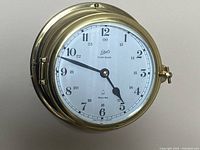 Front view of the brass clock mounted on wall showing the white dial with Schatz logo, Ocean-Quartz marking, and both 12-hour and 24-hour numbering with hour and minute hands.