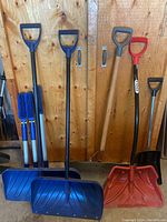 Five shovels standing upright against a wooden backdrop, including two blue snow shovels, one red snow shovel, and two smaller shovels with wooden handles and D-grips.