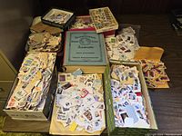 Full lot of the 1947 International Postage Stamp Album surrounded by several boxes and envelopes filled with assorted loose stamps attached to paper scraps.