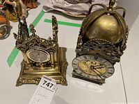 Two brass decorative clocks side by side, showing full view of both, one smaller and gothic-styled, the other larger with dome top
