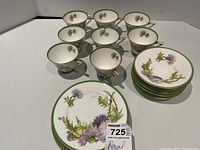 Top view showing eight teacups arranged in two rows and seven saucers and side plates stacked showing the floral pattern and green rims with gold trim for each piece.