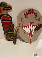Wooden carved bird figure and round painted animal hide shield with feathers, displayed upright.
