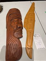 Two carved wood carvings: dark brown human profile panel and light-toned slender stylized carving