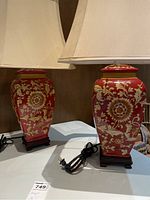 Pair of ceramic base lamps with red and yellow floral design, beige lampshades, on dark square bases, with cords visible, placed on a table
