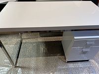 White lacquer rectangular desk top on chrome metal legs beside white lacquer two-drawer file cabinet.
