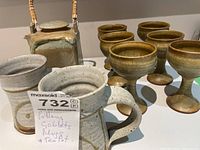 Photo showing six goblets, two mugs, and a square pottery teapot with a woven handle and lid.