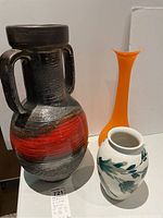 Photo showing all three vases, orange glass vase prominent in back, black and red pottery vase, and white vase with leaf design in front.