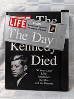 Front cover of the large hardcover book titled 'The Day Kennedy Died' with a black and white image of JFK and a clear removable banner about the included reprint magazine.