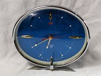 Front view of the retro alarm clock showing the blue oval face with hour markers, white hands, red second hand and chrome base stand.