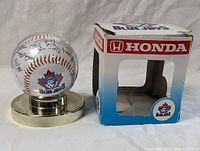 Baseball on metal stand next to original Honda-branded box, showing facsimile autographs and Blue Jays logo on ball