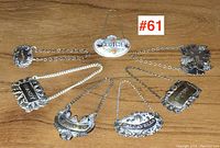 Seven decanter labels displayed on wood surface. Six silver plated, one porcelain with colorful floral design.