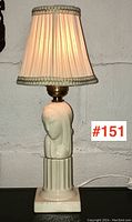 Front view of the lamp with shade lit, showing the pleated cream fabric shade trimmed with decorative metallic braid and the white pottery female bust base on a fluted column pedestal.