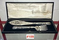 Fish serving set displayed inside original box with black velvet base and cream satin lid lining.