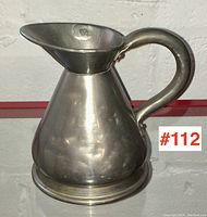 Front view of the pewter pint measure showing conical shape, spout, and handle against a white brick wall.