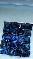 Multiple square-cut natural blue sapphires displayed in a grid pattern inside a jewelry case, showing the vivid blue hues and clarity of the stones.