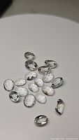 Overhead photo showing 16 clear, oval cut natural crystal quartz gemstones with faceted surfaces, arranged on a plain light background.