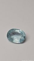 Two photos of a single oval-cut sky blue aquamarine gemstone placed on a neutral background, clearly showing color, cut and clarity.