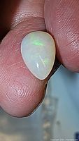 Close-up photo of pear-shaped cabochon cut Ethiopian opal held between fingers showing light green flash on white translucent body.