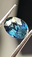 Close-up photo of natural blue sapphire oval cut gemstone held in tweezers, displaying facets and internal inclusions.