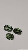 Three oval cut natural green apatite gemstones shown on a neutral background in different lighting angles.