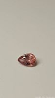 Pear-shaped natural sapphire gemstone with orange-pink color on neutral light background, showing facets and transparency.