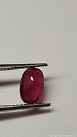 Close-up of a deep red oval cut ruby gemstone held by gem tweezers against a plain background, showing facets and polished surface.