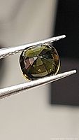 Close-up photo of a cushion-cut olive green tourmaline gemstone held by tweezers showing facets and natural olive color.