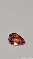 Close-up photo of 0.44ct pear-shaped padparadcha sapphire gemstone on neutral background showing color, cut, and internal features.