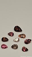 Nine pear-shaped tourmaline gemstones in varying shades of pink and purple, with one almost clear stone, arranged on a neutral background