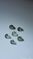 Six pear-shaped faceted natural alexandrite gemstones arranged on a light surface showing subtle color variation.