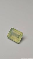 Close-up photo of a rectangular emerald cut mossy green prehnite gemstone with visible rutile and black tourmaline inclusions inside.