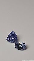 Photo showing the two natural Tanzanite gemstones: one trillion cut and one oval cut, both purple blue in color, placed on a flat surface.