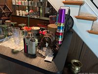 Multi-colored aluminum cups stacked, martini glasses, flask, coasters, and ceramic barrel on stand arranged on table near staircase.