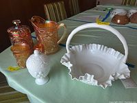 Milk glass basket with handle and ruffled edge placed alongside Avon lidded dish and carnival glass items on table