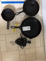 Three cast iron frying pans and four trivets laid out on the floor for display.
